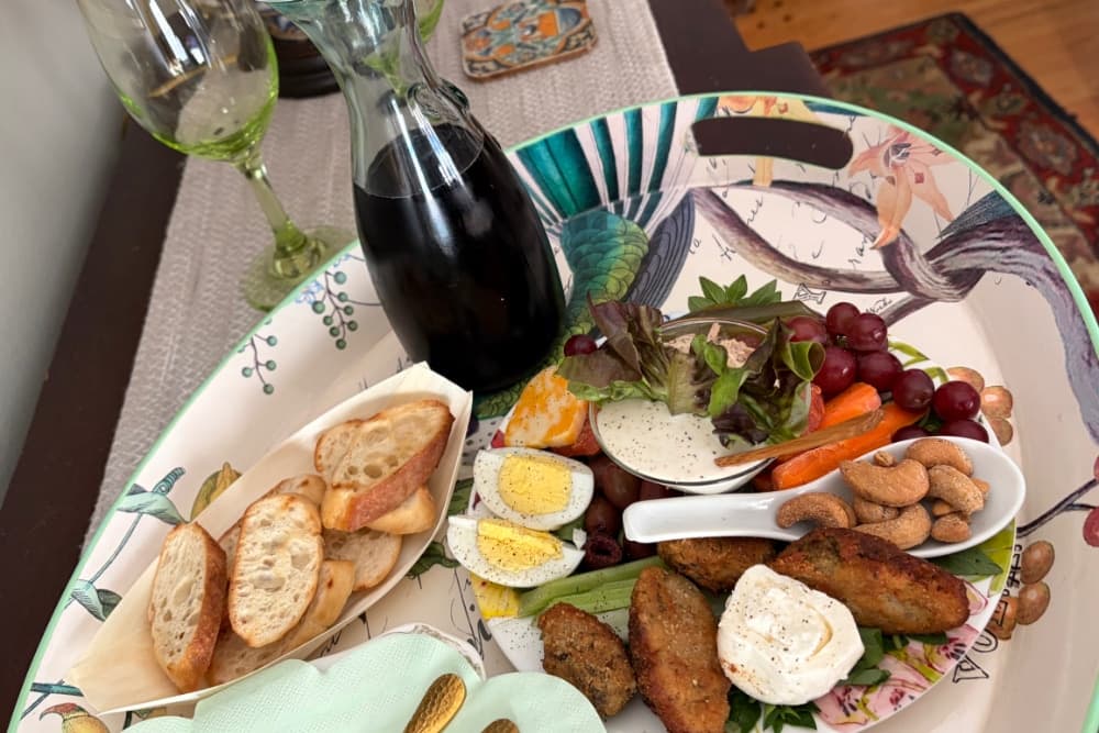 A decorative tray filled with assorted snacks including bread, salad, eggs, grapes, carrots, nuts, and a dark beverage.