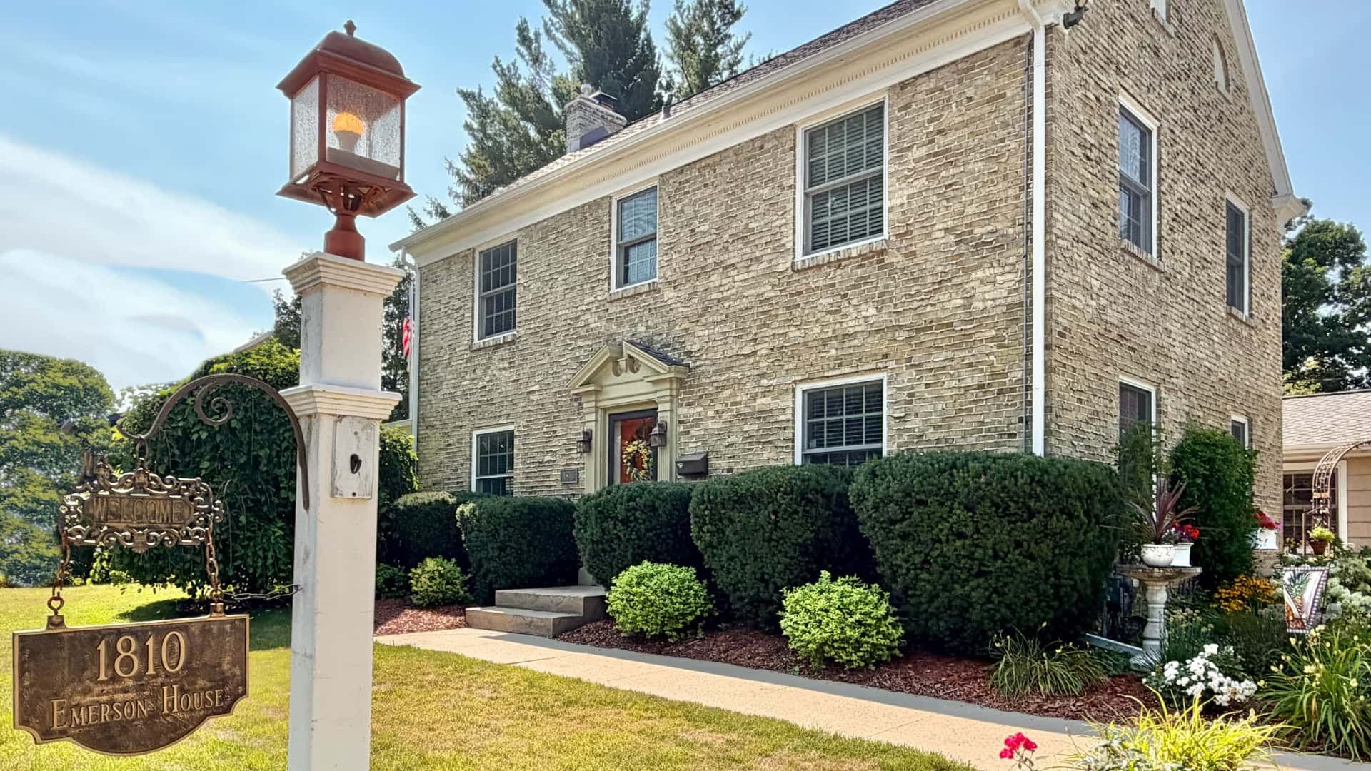 Two-story brick house with a sign reading "1810 Emerson House" and lush greenery in front.