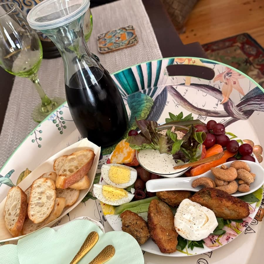 A colorful platter of appetizers including fried food, vegetables, fruit, and a glass bottle of dark liquid, accompanied by bread and utensils.