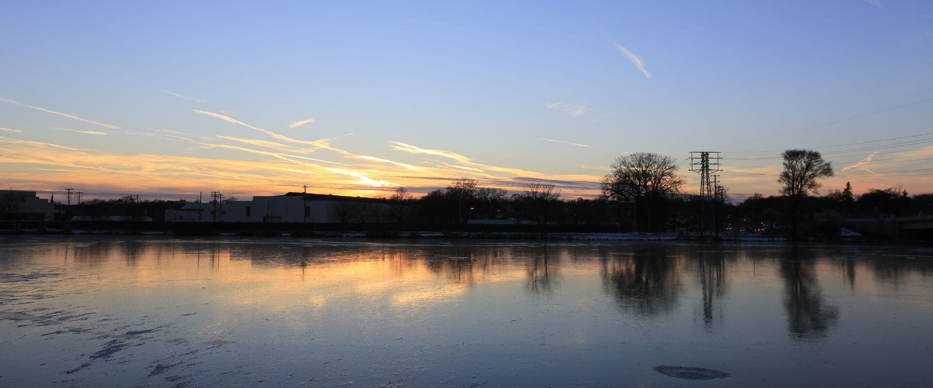Sunset over a reflective lake with silhouettes of trees and buildings.