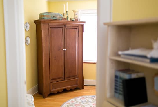 A wooden armoire stands in a softly lit, yellow-walled room with decorative plates and shelves nearby.