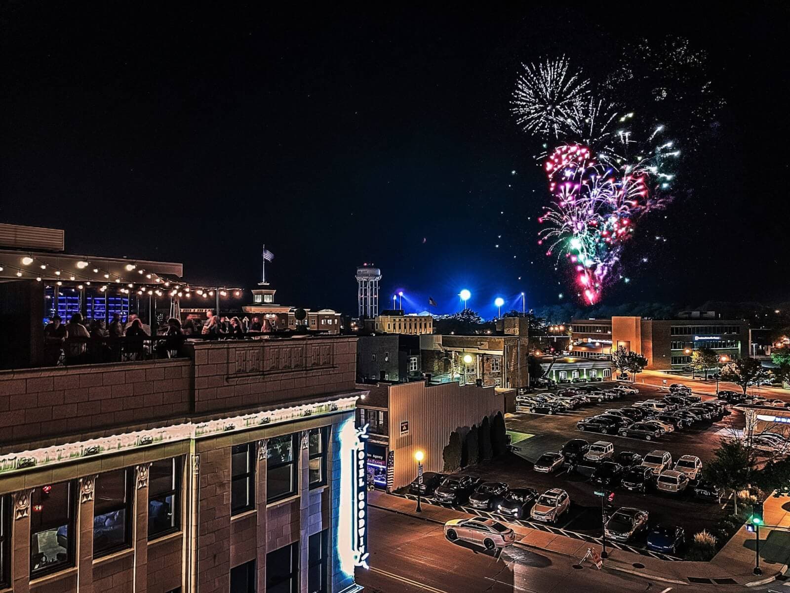 A nighttime urban skyline view with fireworks bursting above a busy street and rooftop crowd.
