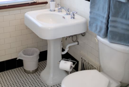 A small bathroom featuring a pedestal sink, toilet, and wastebasket on a tiled floor.