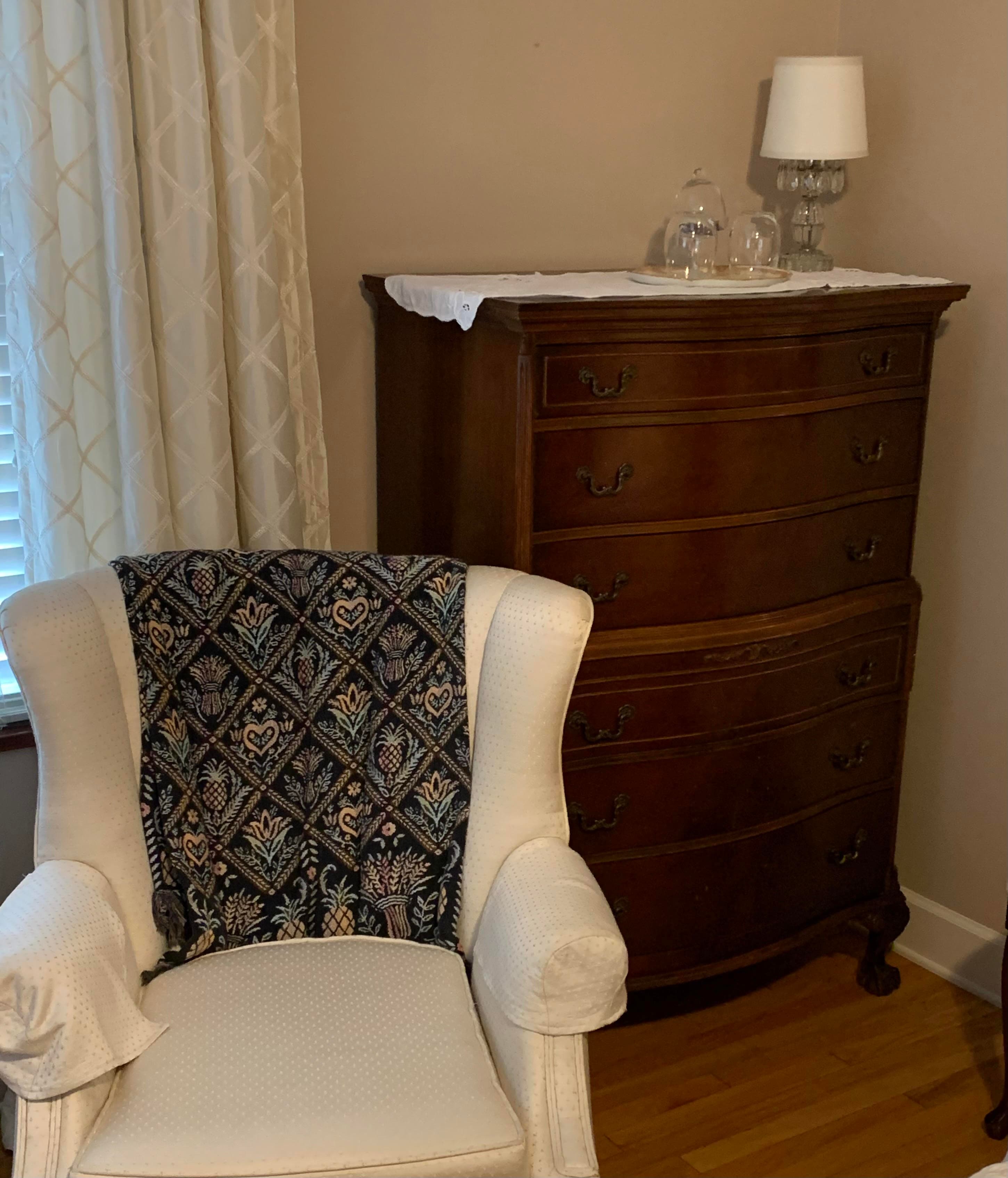 A cozy room corner featuring a white armchair draped with a patterned blanket beside a wooden dresser and a lamp.