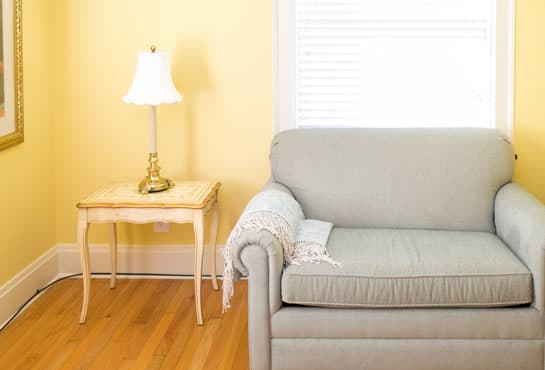 A light gray couch and a small side table with a lamp against a yellow wall.