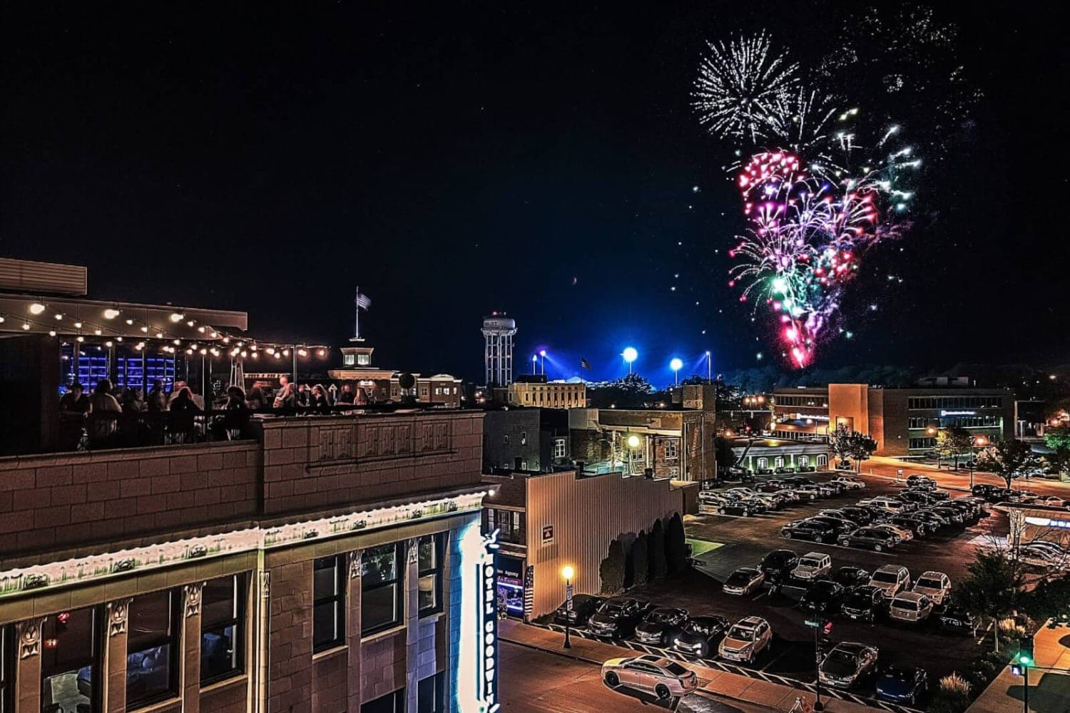 Fireworks illuminate the night sky above a bustling cityscape with a rooftop gathering.
