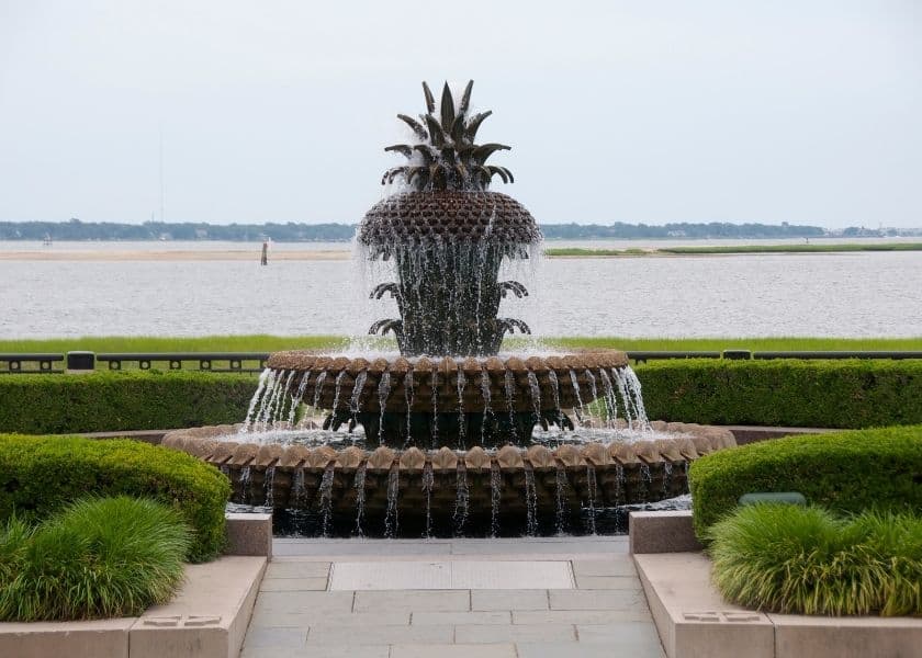 A pineapple-shaped fountain surrounded by lush greenery and a serene waterfront view.