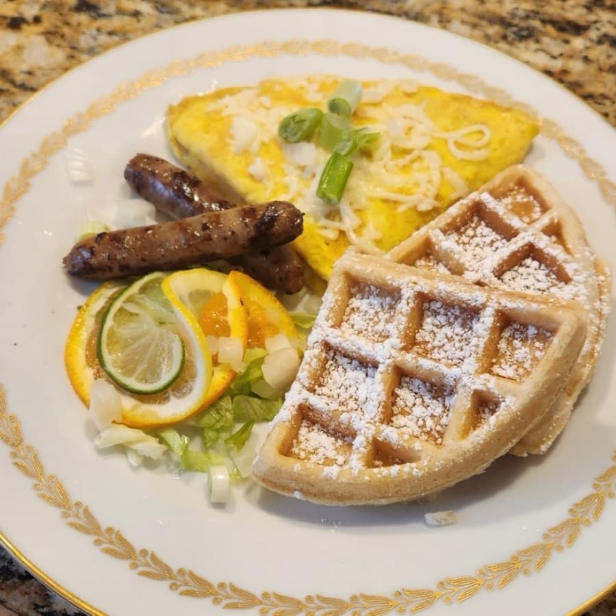 A plate with an omelet topped with green onions, two sausage links, waffles dusted with powdered sugar, and a side of salad with citrus slices.