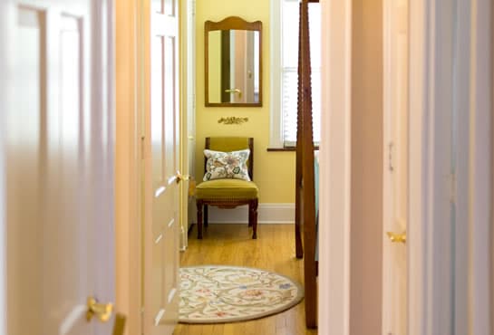 A narrow hallway featuring a decorative rug, a chair with a patterned cushion, and a wall mirror.